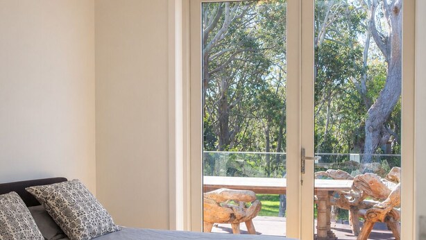 A bedroom with a large bed, a view of the outdoors through a glass door, and a wooden floor. - All The Views - Ocean & Bush with Heated Infinity Lap and Play Pool (Otford)