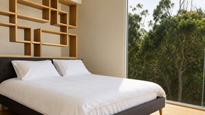 A modern bedroom with a large bed, a dark headboard, and a view of trees through a large window. - All The Views - Ocean & Bush with Heated Infinity Lap and Play Pool (Otford)