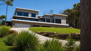 A modern two-story house with a garage, surrounded by a well-maintained lawn and native vegetation. - All The Views - Ocean & Bush with Heated Infinity Lap and Play Pool (Otford)