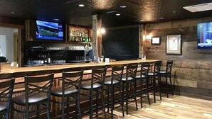 A bar area with a long counter, multiple stools, a television screen, and a wall-mounted shelf with bottles. - Shady Creek Cabin in Connestee Falls with stream (Brevard)