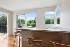 A modern kitchen with a large island, two wicker chairs, and a view of greenery through large windows.