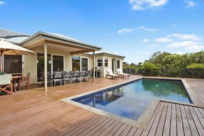Ein Schwimmbecken mit Holzterrasse, Gartenmöbeln und einer überdachten Terrasse.