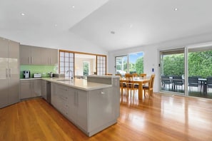 A modern kitchen with a central island, wooden flooring, and a dining area with a view of greenery.