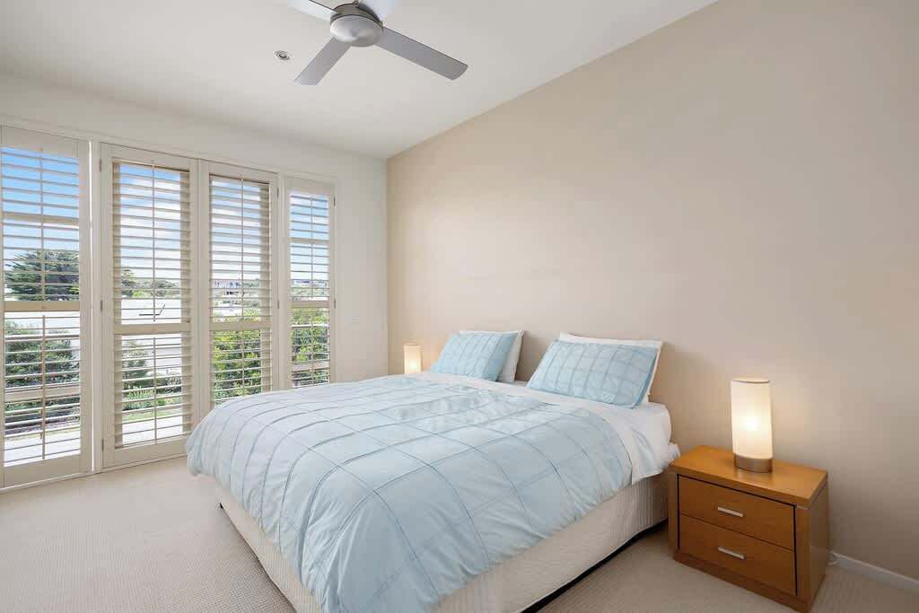 Une chambre à coucher comprenant un lit, deux tables de chevet, un ventilateur de plafond et une vue dégagée sur l’extérieur grâce à de grandes fenêtres.