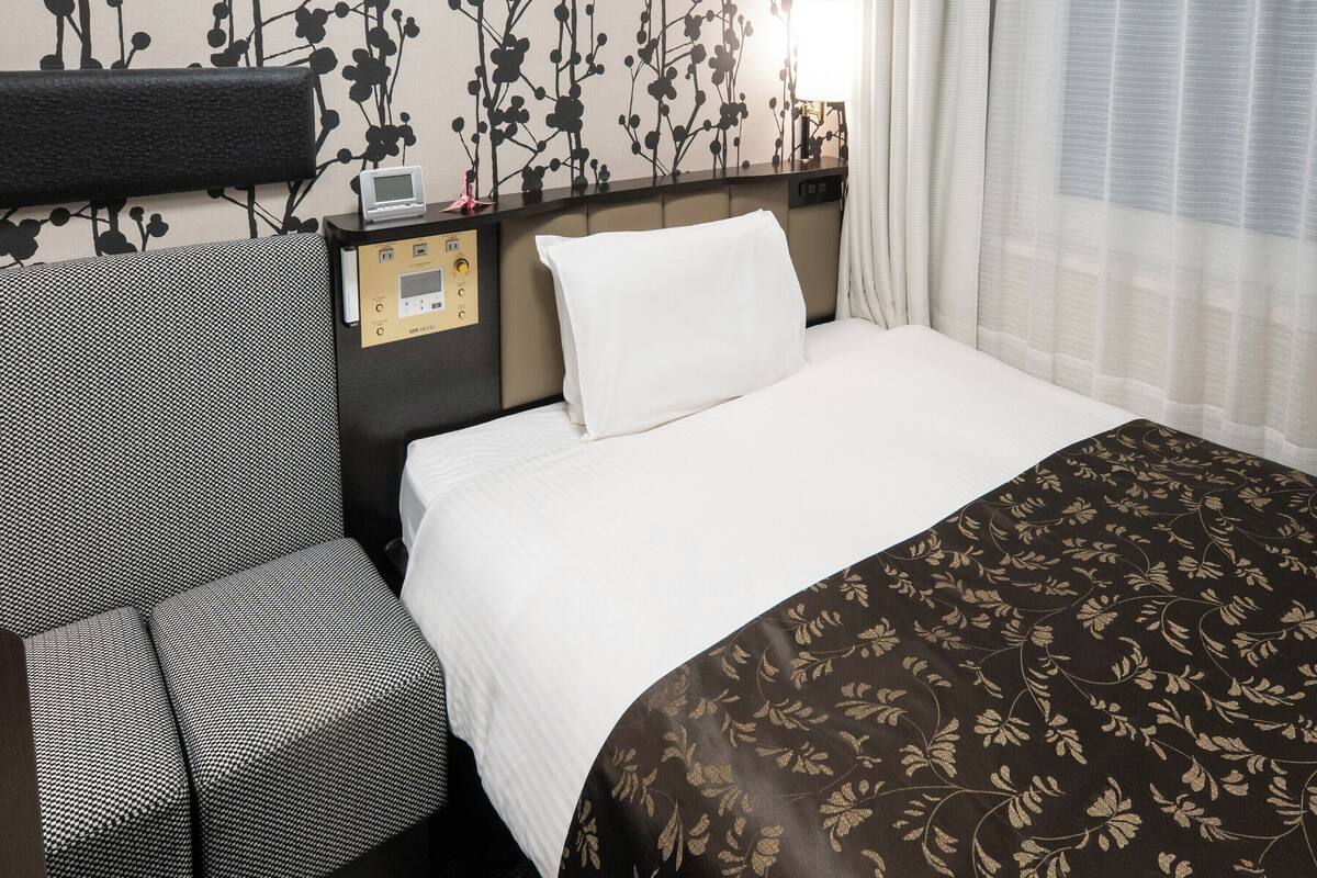 Hotelkamer met een bed, bedieningspaneel, stoel en raam met gordijnen.