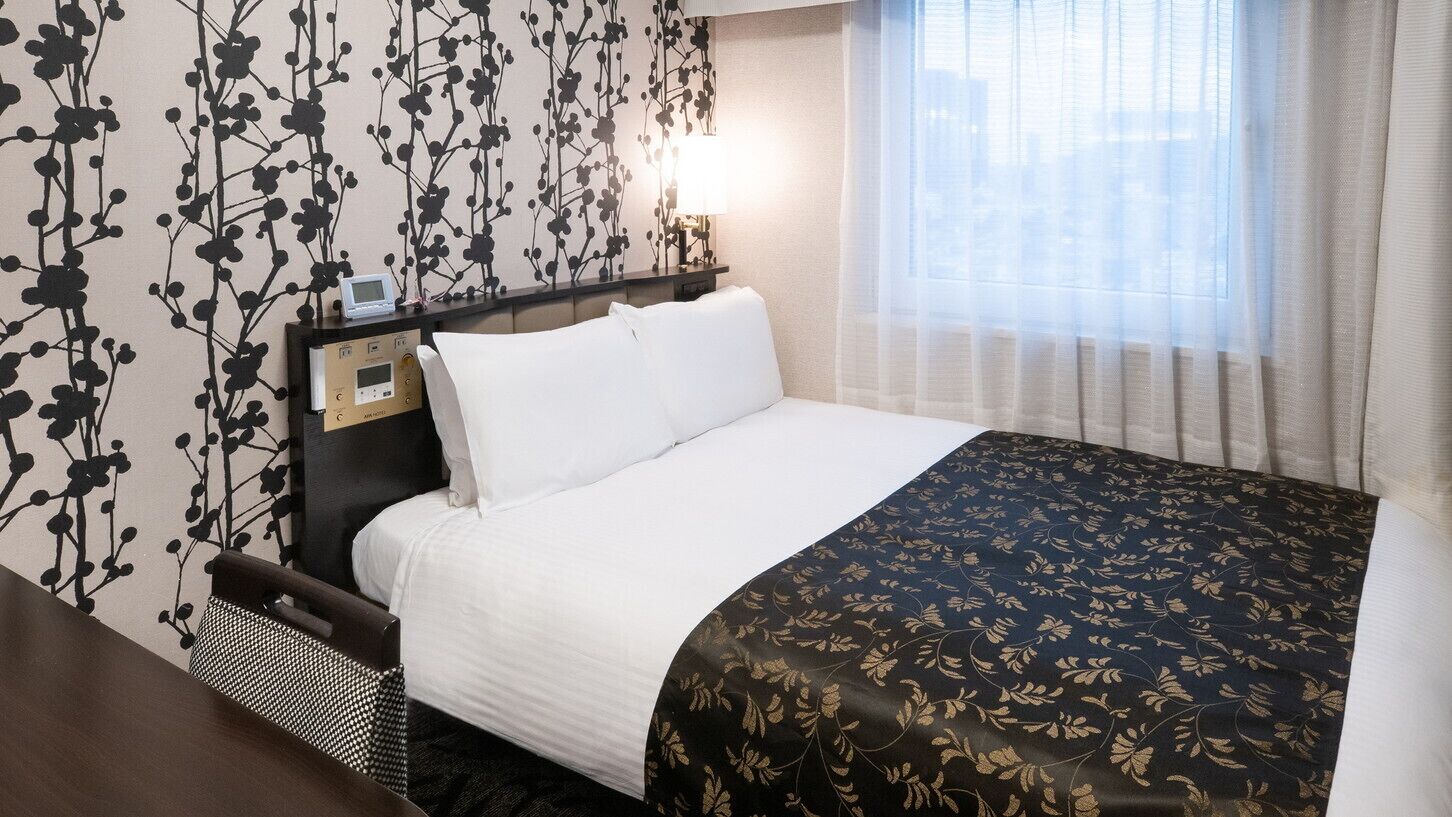 A hotel room with a bed, a bedside table with a clock, a window with sheer curtains, and a wall with a black floral pattern.