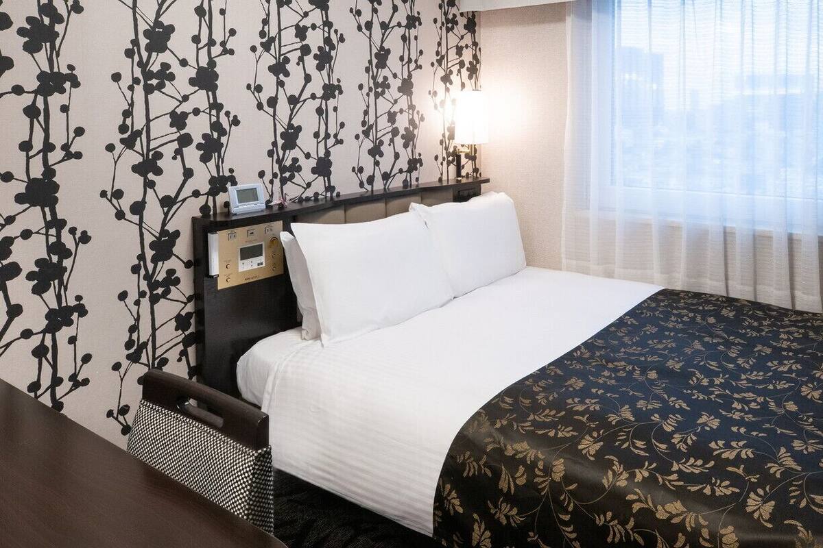 Een hotelkamer met een bed, een nachtkastje met een klok, een raam met dunne gordijnen en een muur met een zwart bloemenpatroon.