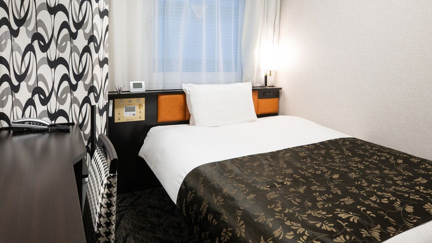 A hotel room with a bed, a bedside table with a phone and a digital clock, a headboard, a window with curtains, and a wall with a textured pattern.