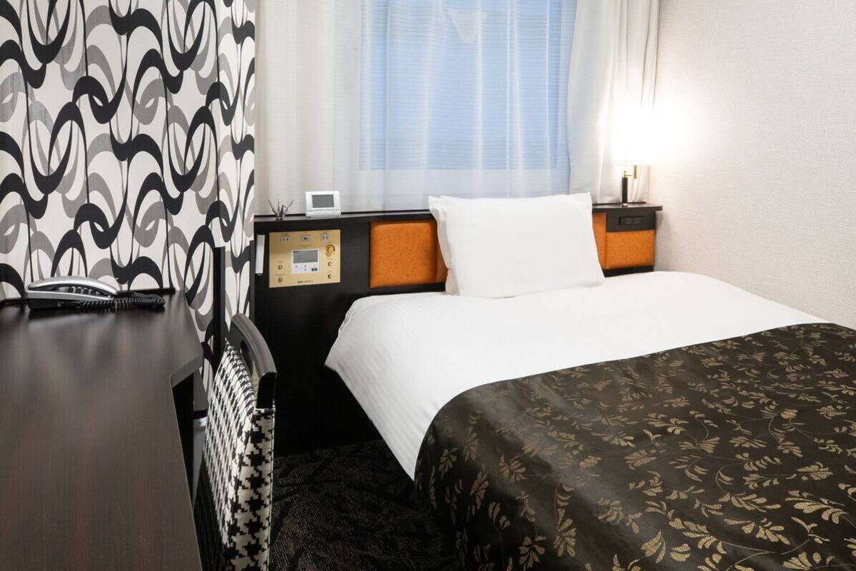 Hotelkamer met een bed, nachtkastje met telefoon en digitale klok, hoofdeinde, raam met gordijnen en een wand met een structuur.