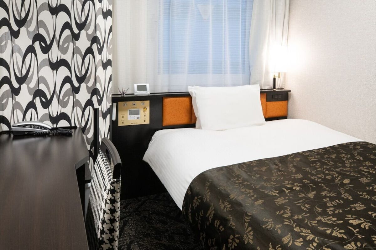 Habitación de hotel con cama, mesita de noche con teléfono y reloj digital, cabecero, ventana con cortinas y pared con textura.