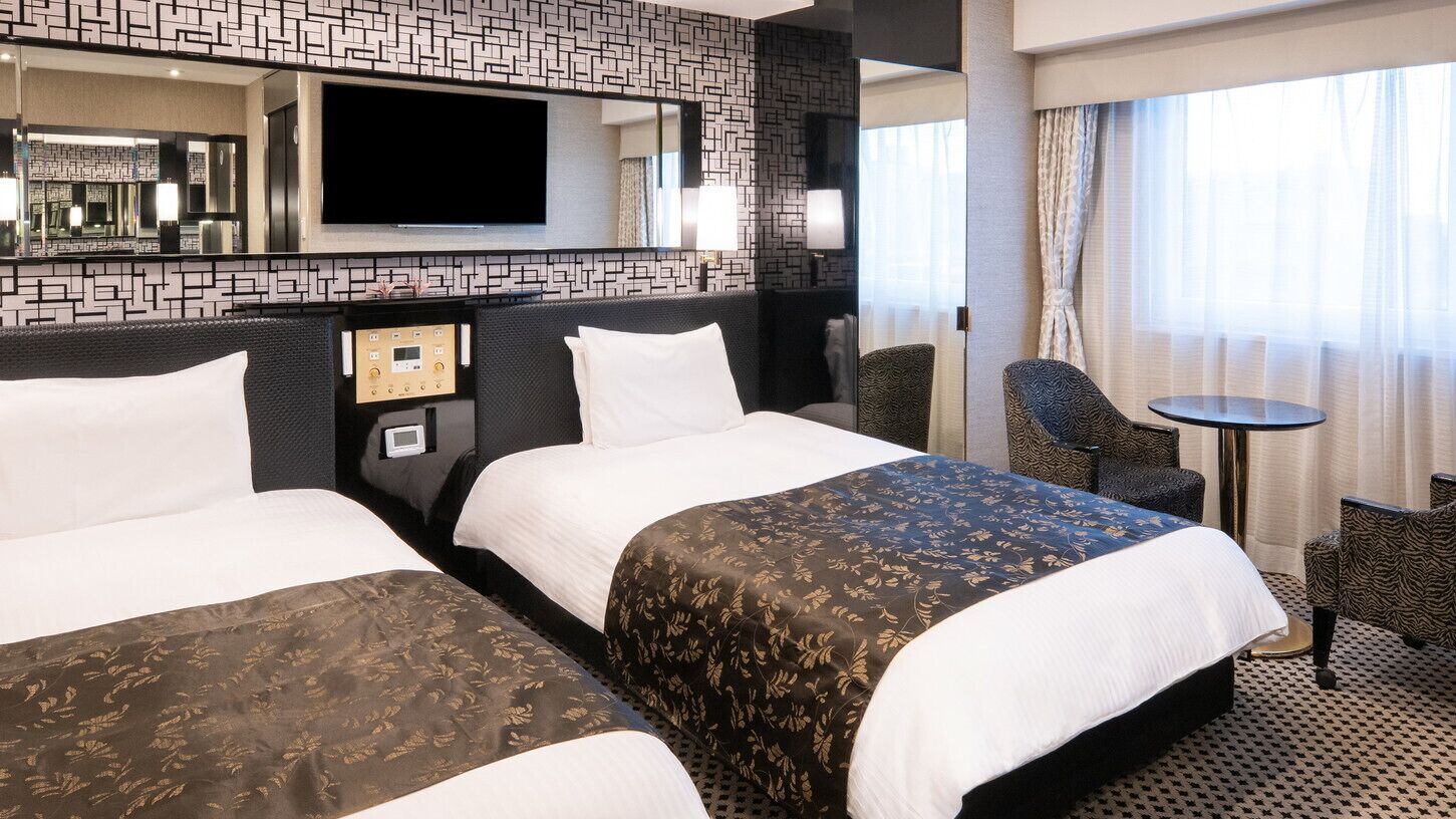 A hotel room with two beds, a TV, a control panel, a small table, and a chair.