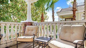 A covered outdoor seating area with two chairs and a table, surrounded by a white balustrade and palm trees. - Beautiful Condo with Golf Course Views, Near Attractions (Kissimmee)