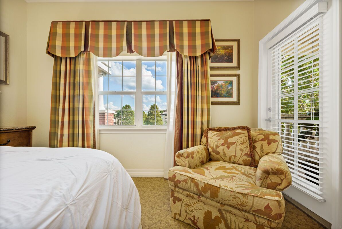 A bedroom with a bed, a floral-patterned armchair, a window with curtains, and a picture frame on the wall.