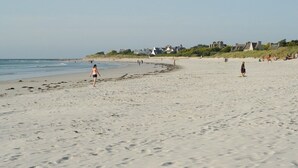 Une vaste plage de sable où l’on voit quelques personnes se promener et se tenir debout. On aperçoit des maisons au loin, le long du rivage.