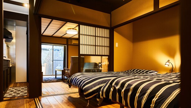 Un dormitorio tradicional japonés con piso de madera, una cama con ropa de cama a rayas y una puerta corredera que da a un balcón.