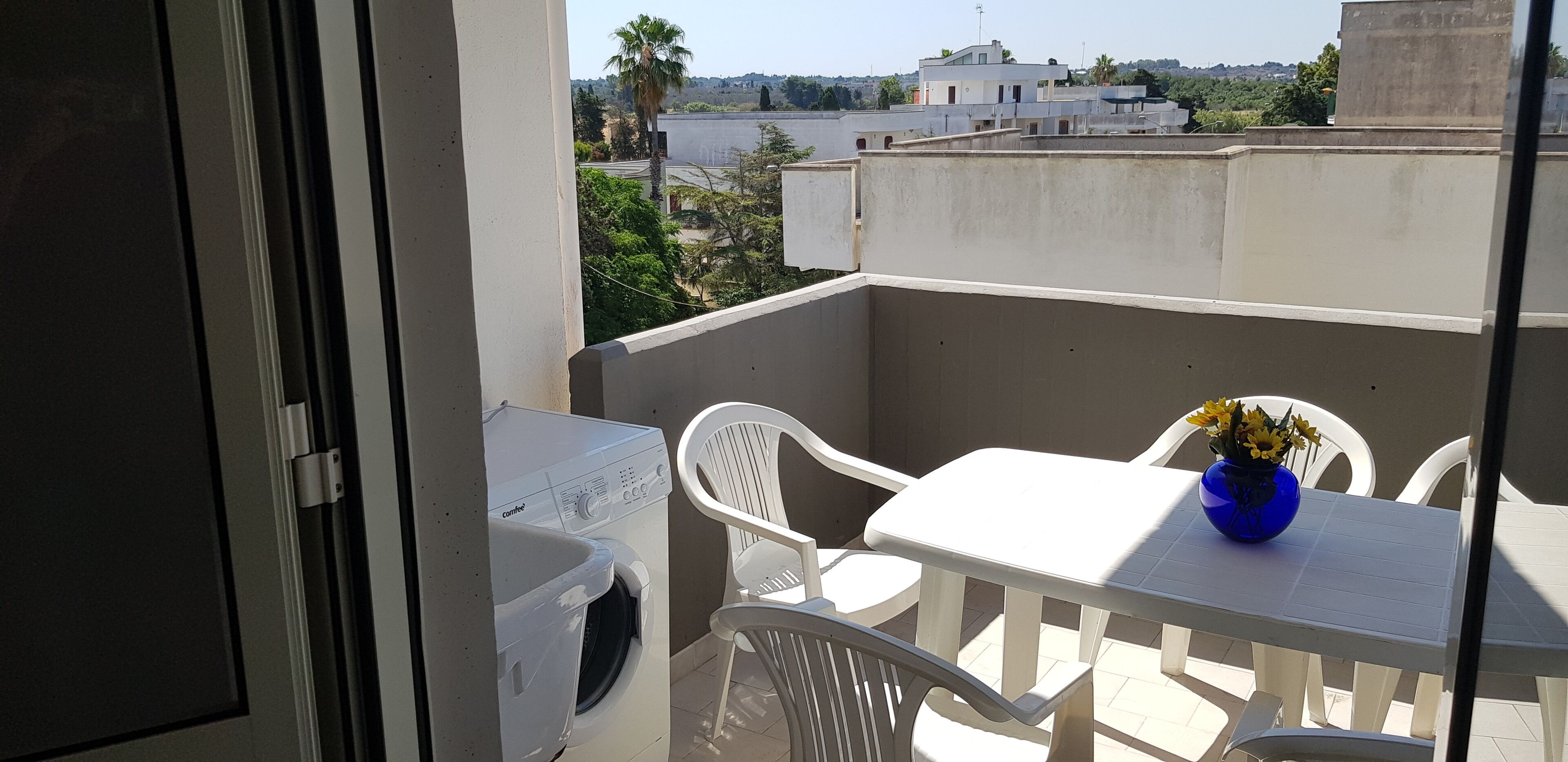 A balcony with a washing machine, white plastic chairs, a table, and a vase with flowers.