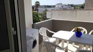 A balcony with a washing machine, white plastic chairs, a table, and a vase with flowers.