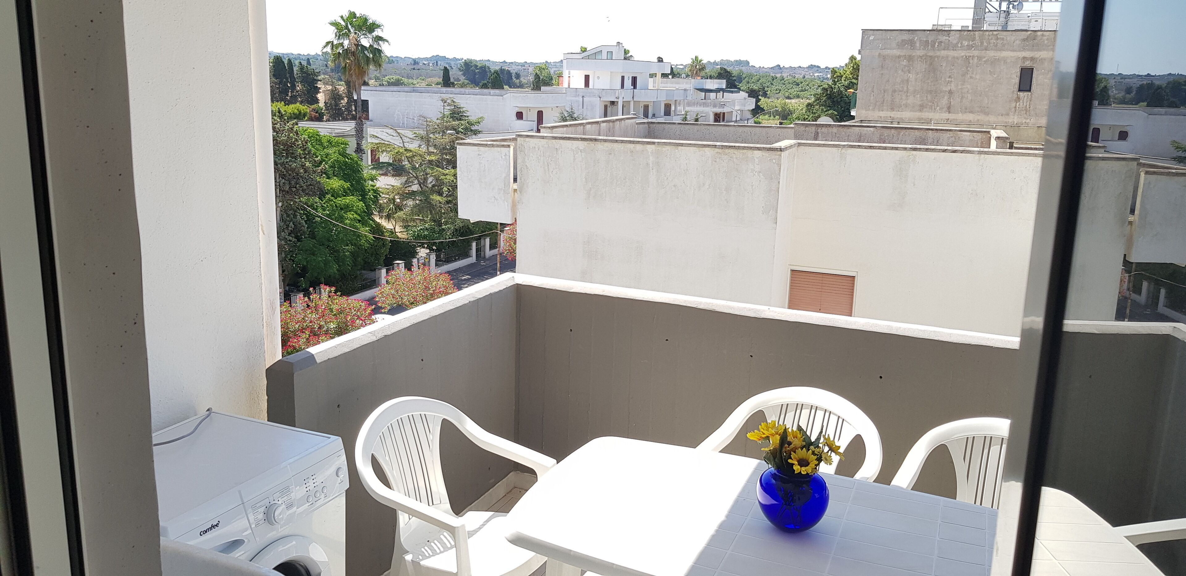 Un balcón con sillas de plástico blanco, una mesa y un jarrón azul con flores.