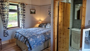 A bedroom with a bed, a window with curtains, a bedside lamp, and a wooden door leading to a bathroom with a sink and mirror. - Kate's Cottage (Valentia Island)