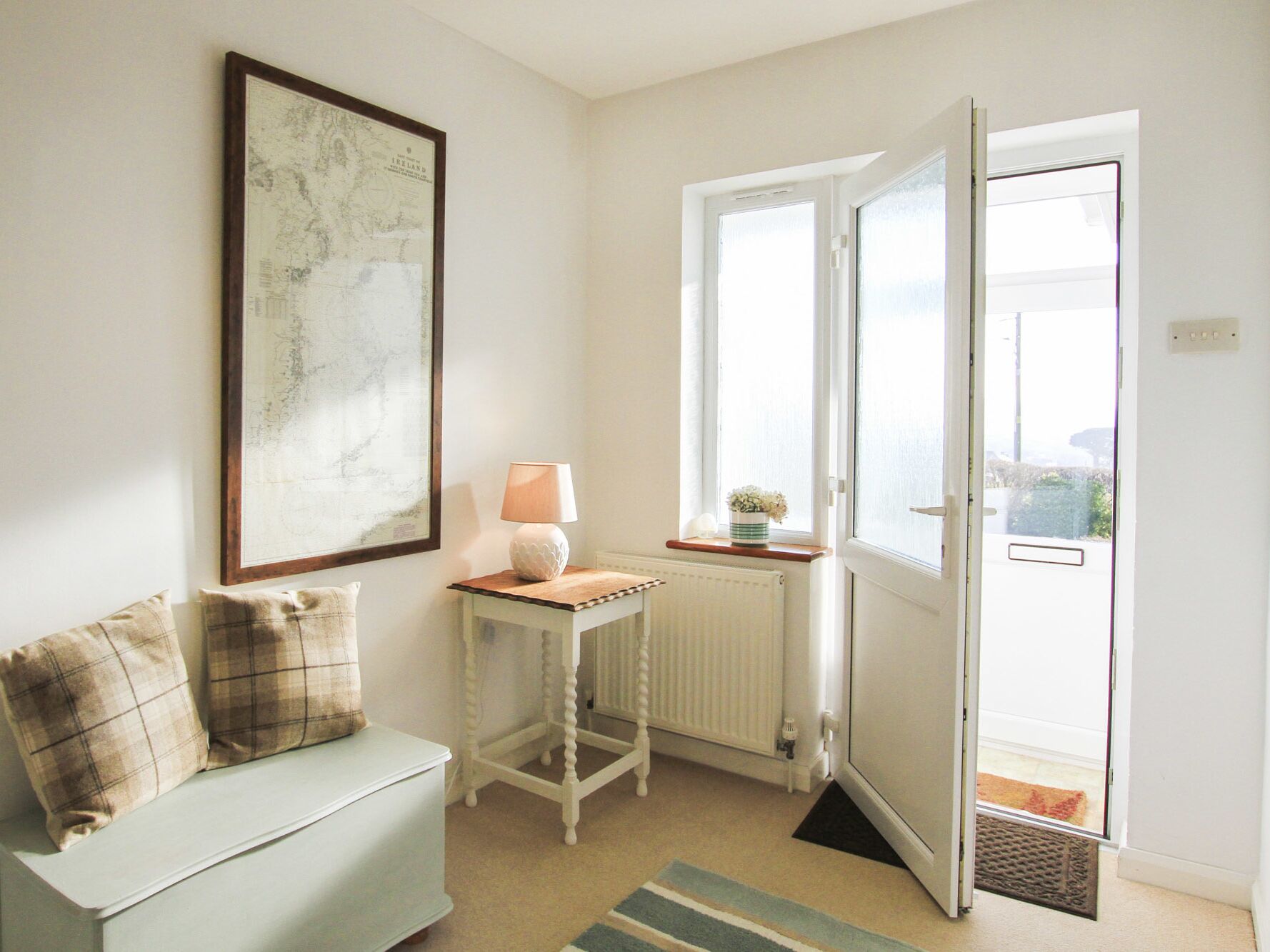 A room with a white radiator, a white table with a lamp, a white sofa with plaid cushions, and a large map on the wall.