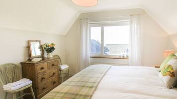A bedroom with a large bed, a wooden dresser, a chair, a window with a view, and a lamp.