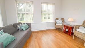 Una habitación con un sofá gris, dos sillones de mimbre, una mesita con una lámpara y ventanas amplias con persianas.