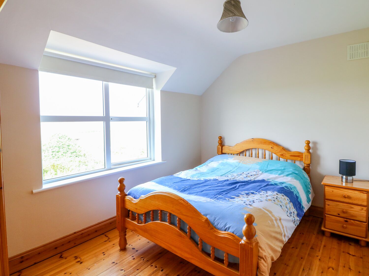 A bedroom with a wooden bed, a nightstand, a lamp, and a window with a view of greenery.