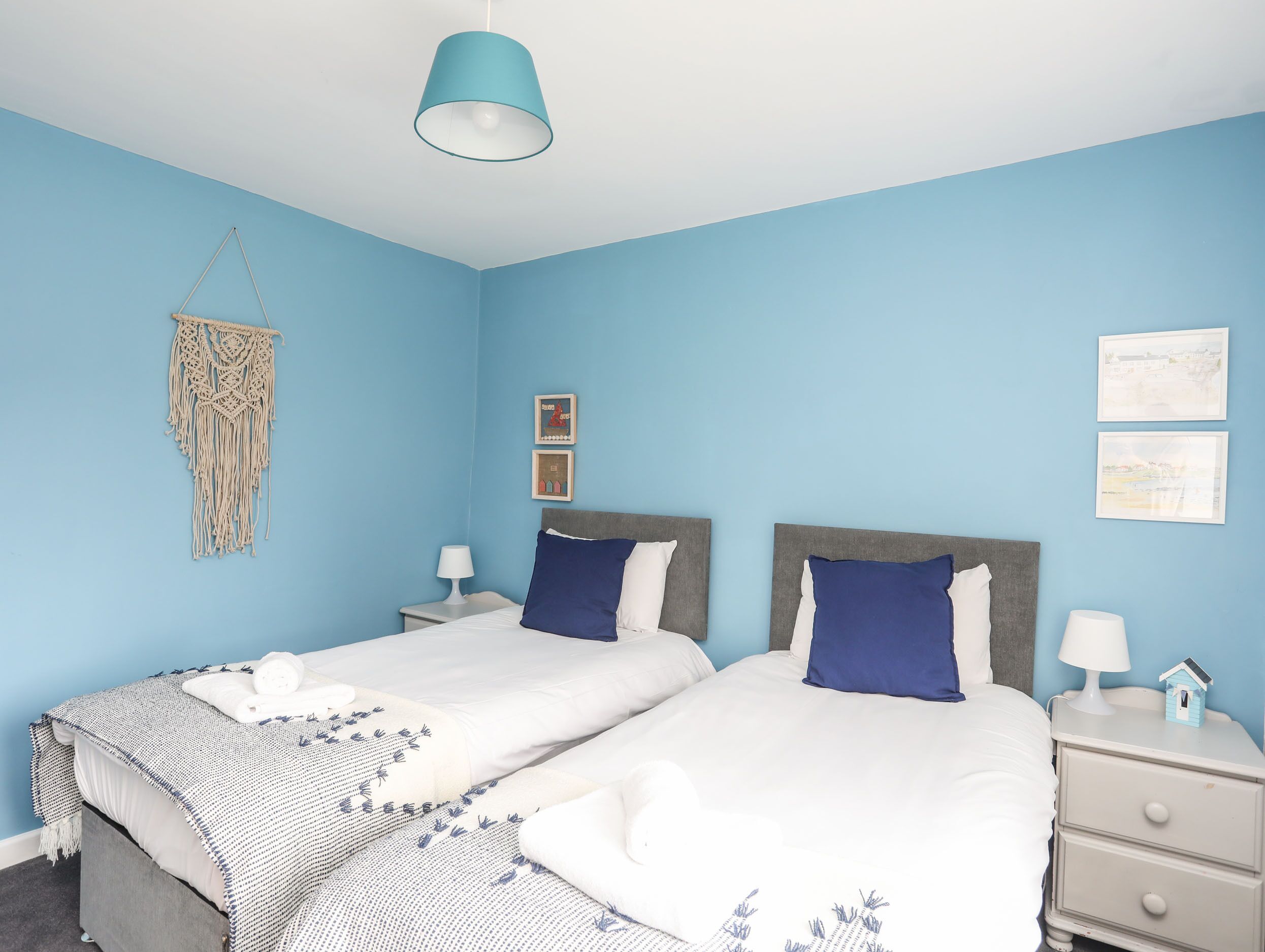 A bedroom with two beds, a hanging macrame decoration, and framed pictures on the wall.