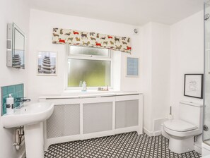 A bathroom with a white sink, toilet, and shower. There is a window with a patterned curtain and a framed picture on the wall. - Gorwel (Moelfre)