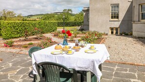 A table set outdoors with a white tablecloth, surrounded by a garden with hedges and flowers, and a building in the background. - The Lodge at Raheengraney House (Raheengraney)