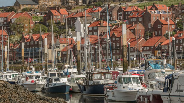 A marina with boats docked, surrounded by traditional red-roofed buildings and a historic stone structure in the background.