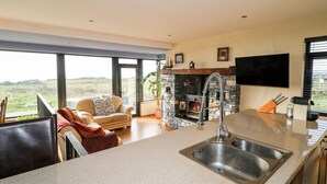 A modern kitchen with a stone fireplace, large windows, and a dining area with a view. - Ocean Sail House (Maghery)