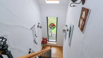 A narrow stairway with a wooden handrail, a small window with a stained glass design, and wall-mounted decor including a candle holder and a framed picture.