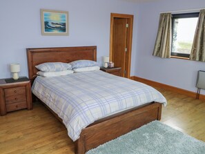 A bedroom with a wooden bed, a nightstand with a lamp, a dresser, a painting on the wall, and a window with curtains. - Ocean Sail House (Maghery)