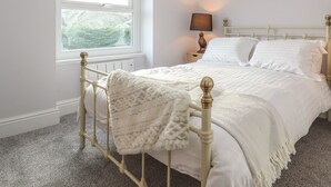 Un lit bien fait, avec une couette blanche et un plaid, une table de chevet avec une lampe, et une fenêtre donnant sur un paysage verdoyant.