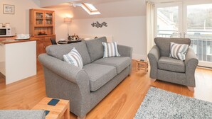 A modern living room with a grey sofa, a grey armchair, and a wooden coffee table. There is a dining area with a table and chairs, and a kitchen area with a microwave and cabinets. - 42 Dart Marina (Dartmouth)