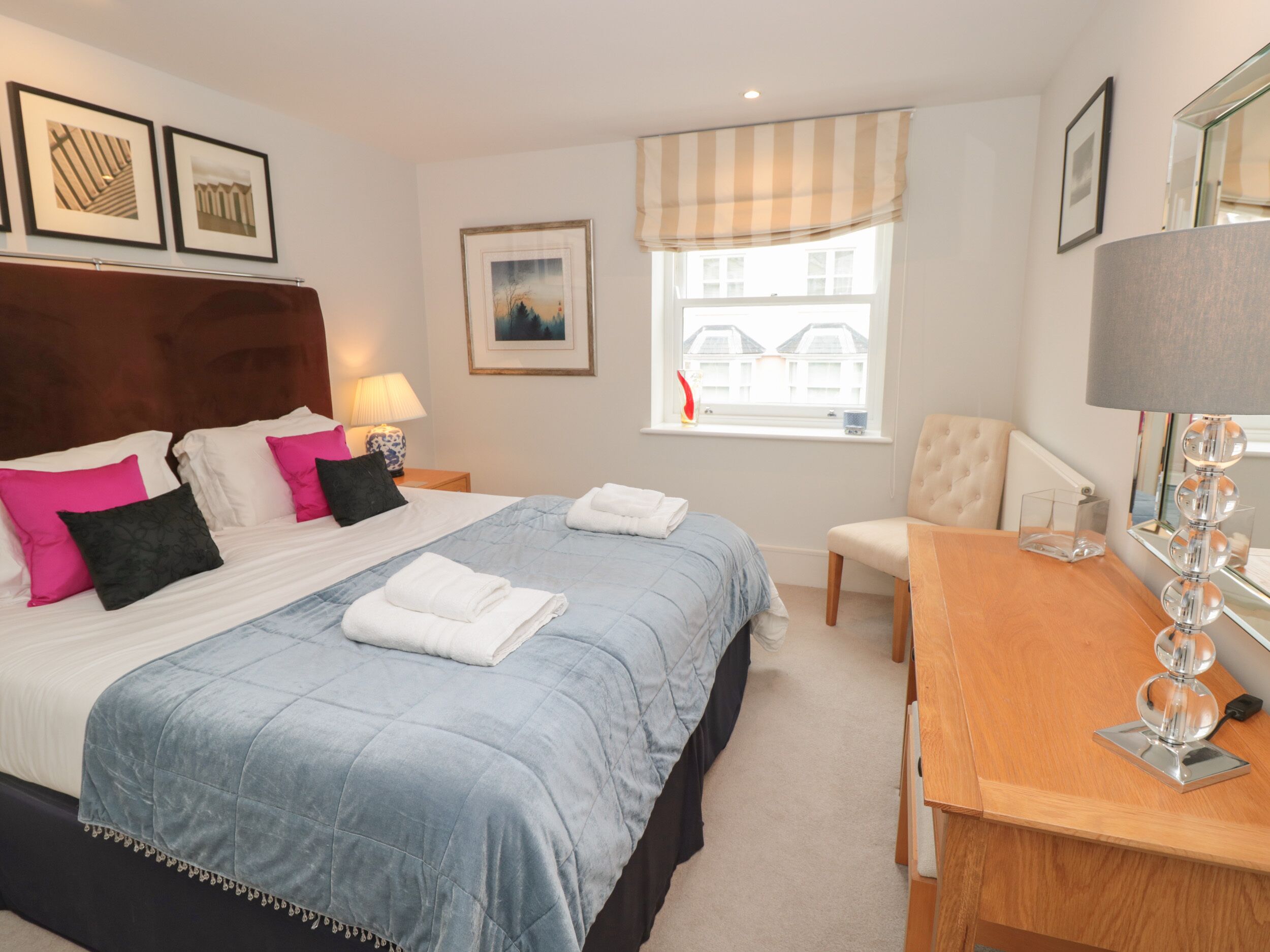 A neatly arranged bedroom with a bed, bedside tables, a chair, a desk, and a window with a view of buildings.