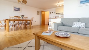 Una sala de estar moderna con un sofá gris, una mesa de comedor de madera y sillas. Hay una zona de cocina con armarios blancos y una pequeña isla.