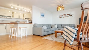 Una sala de estar moderna con un sofá gris, suelo de madera y un comedor con armarios blancos y taburetes.