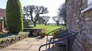 A stone patio with a metal chair, potted plants, and a hedge. - Cartwheel Cottage (York)