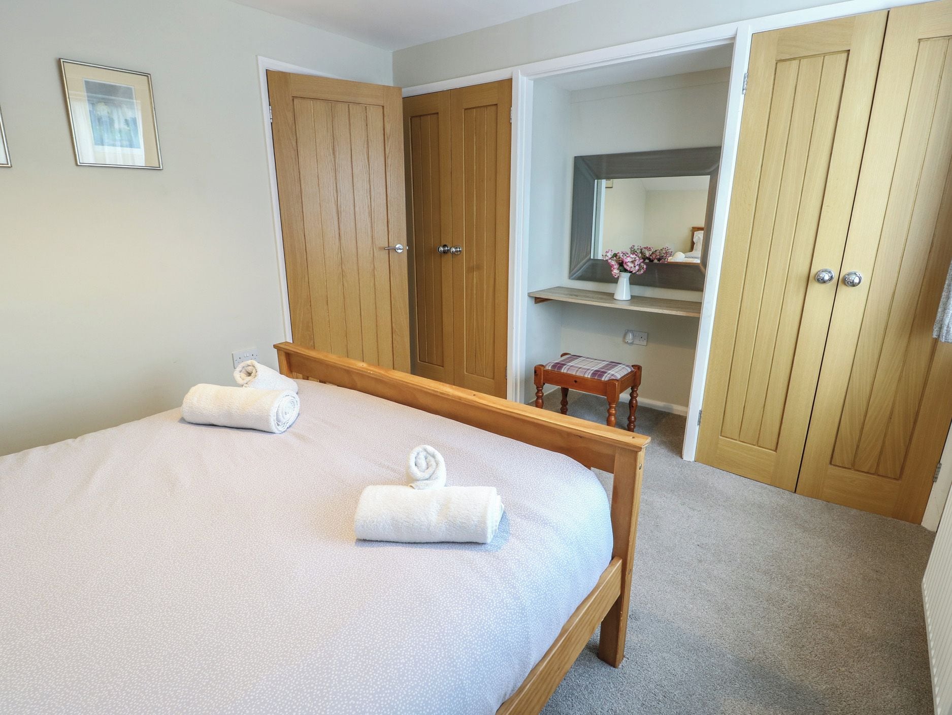 Un dormitorio con una cama, dos puertas de madera, una mesita con un florero y un espejo.
