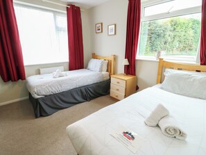 A hotel room with two beds, a nightstand, a lamp, and a window with curtains. - Awelon (Colwyn Bay)