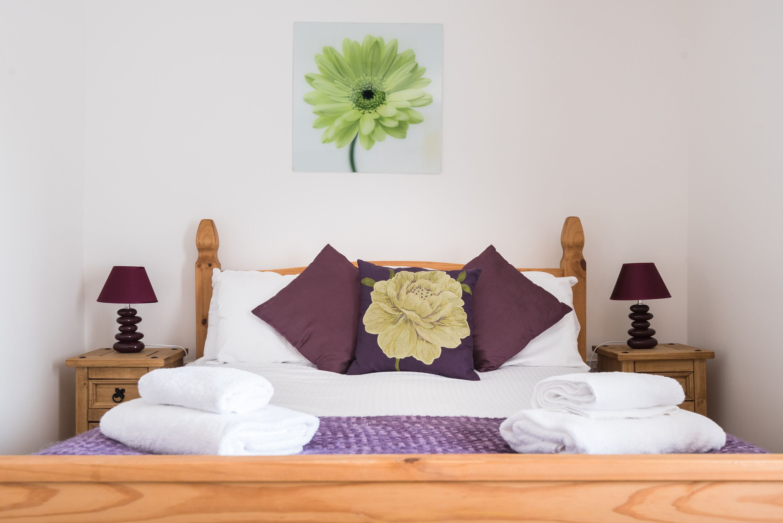 A neatly made bed with white linens, a purple throw, and a large floral pillow. There are two wooden nightstands with lamps and folded towels.