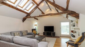 Een ruime woonkamer met een hoge plafon, zichtbare houten balken, een grijze bank, een houten salontafel, een televisie en een boekenkast.