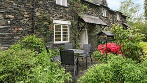 A stone building with a small patio area featuring a table and chairs, surrounded by lush greenery and vibrant flowers.