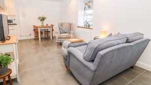 Ein modernes Wohnzimmer mit grauem Sofa, einem Esstisch mit Stühlen, einer Küchenzeile mit Mikrowelle und einem Fenster mit Vorhängen.