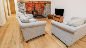 A cozy living room with a stone fireplace, two grey sofas, a wooden TV stand, and a wooden floor. - Tŷ'n Lôn Bach (Caernarfon)