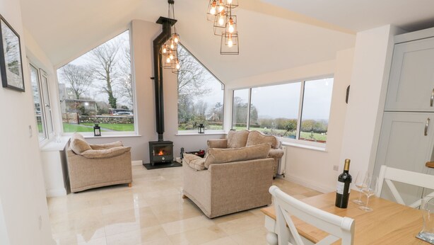 A spacious living area with a fireplace, two sofas, a dining table with chairs, and large windows offering a view of the outdoors. - Tŷ'n Lôn Bach (Caernarfon)