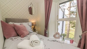 A bedroom with a bed featuring a deer print bedspread, a nightstand with a lamp, and a window with a view of trees. - Bank End Cottage (Carnforth)