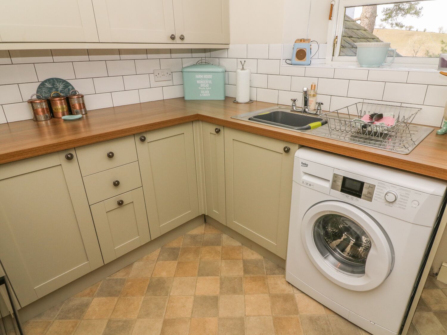 Une cuisine avec des meubles de couleur beige, un lave-linge, un évier avec un égouttoir et une fenêtre donnant sur des arbres.